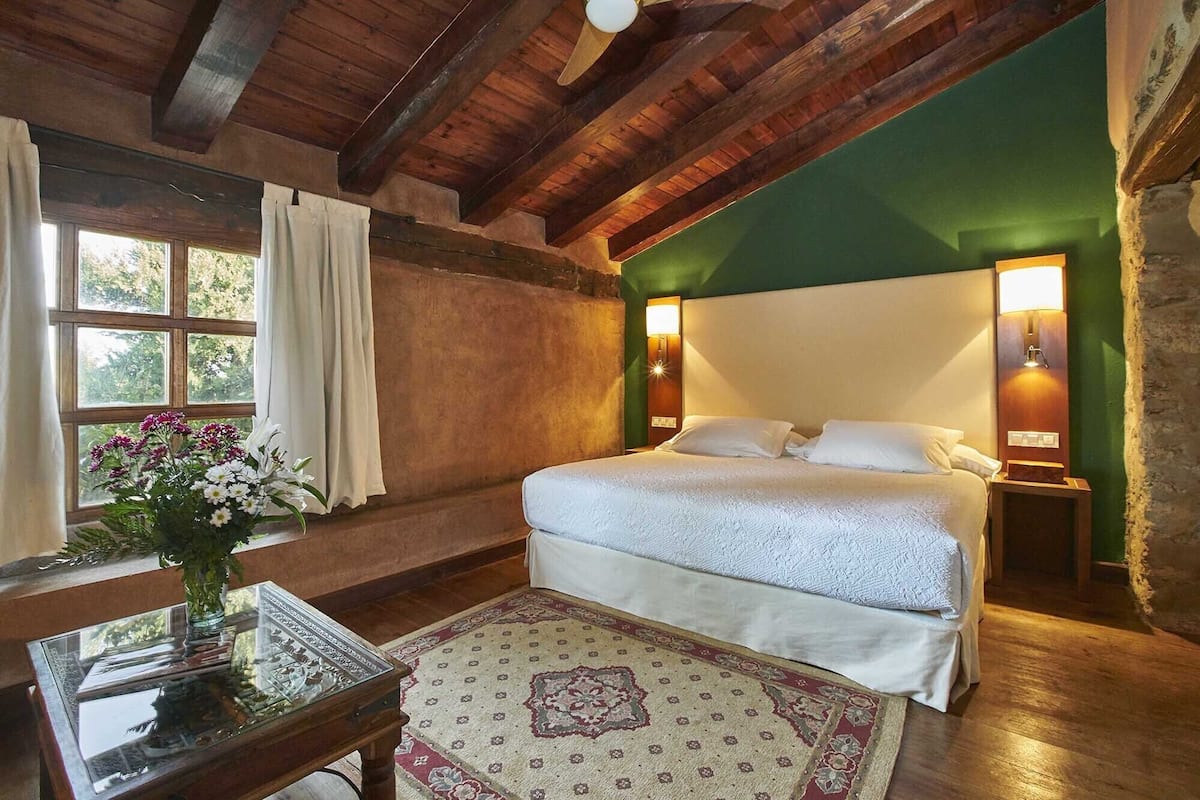 A cozy bedroom with a wooden ceiling, a large bed with white linens, a nightstand with a lamp, and a vase of flowers on a table.