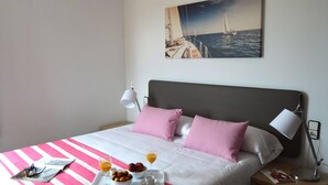 A neatly made bed with a pink and white striped bedspread, a tray with breakfast items, and a painting of sailboats on the wall. - Pierre & Vacances Comarruga (El Vendrell)
