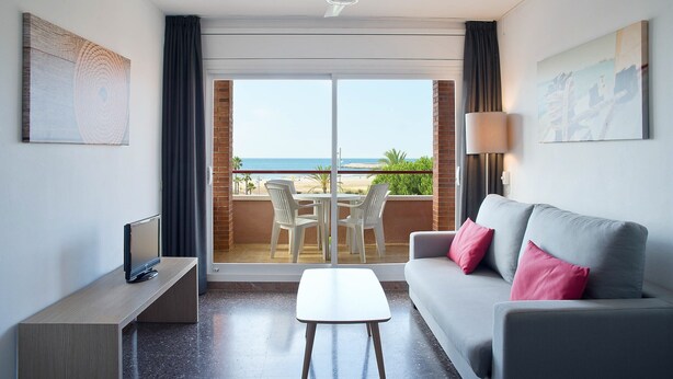 A modern hotel room with a balcony, a grey sofa, a TV, and a dining table with chairs. - Pierre & Vacances Comarruga (El Vendrell)
