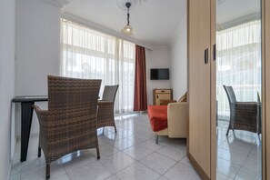 A hotel room with a dining area, two wicker chairs, a sofa, a TV, and a large window with curtains.