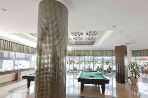 Una mesa de billar en un espacio interior bien iluminado, con ventanas amplias y un techo decorativo.