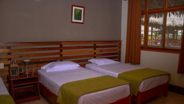 A room with two beds, a wooden headboard, a nightstand with a lamp, and a window with curtains. - Heliconia Amazon River Lodge (Indiana)