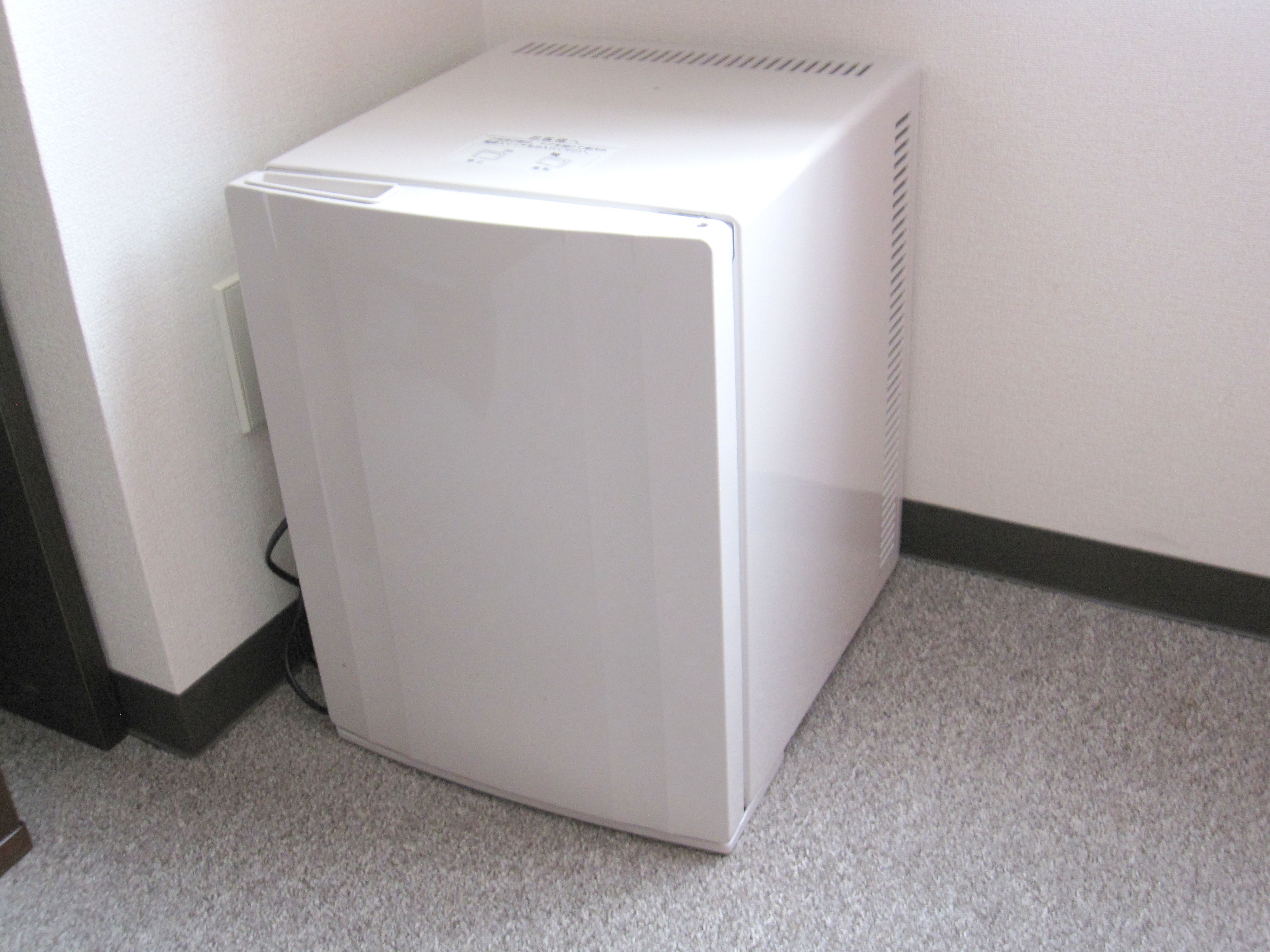 Un refrigerador blanco y compacto en un rincón de una habitación con piso alfombrado y pared blanca.