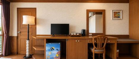 Habitación de hotel con escritorio de madera, televisor, silla, lámpara de escritorio, cafetera y un refrigerador pequeño.