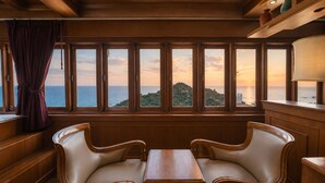 A room with a large window overlooking a coastal view, two leather chairs, a wooden table, and a curtain.