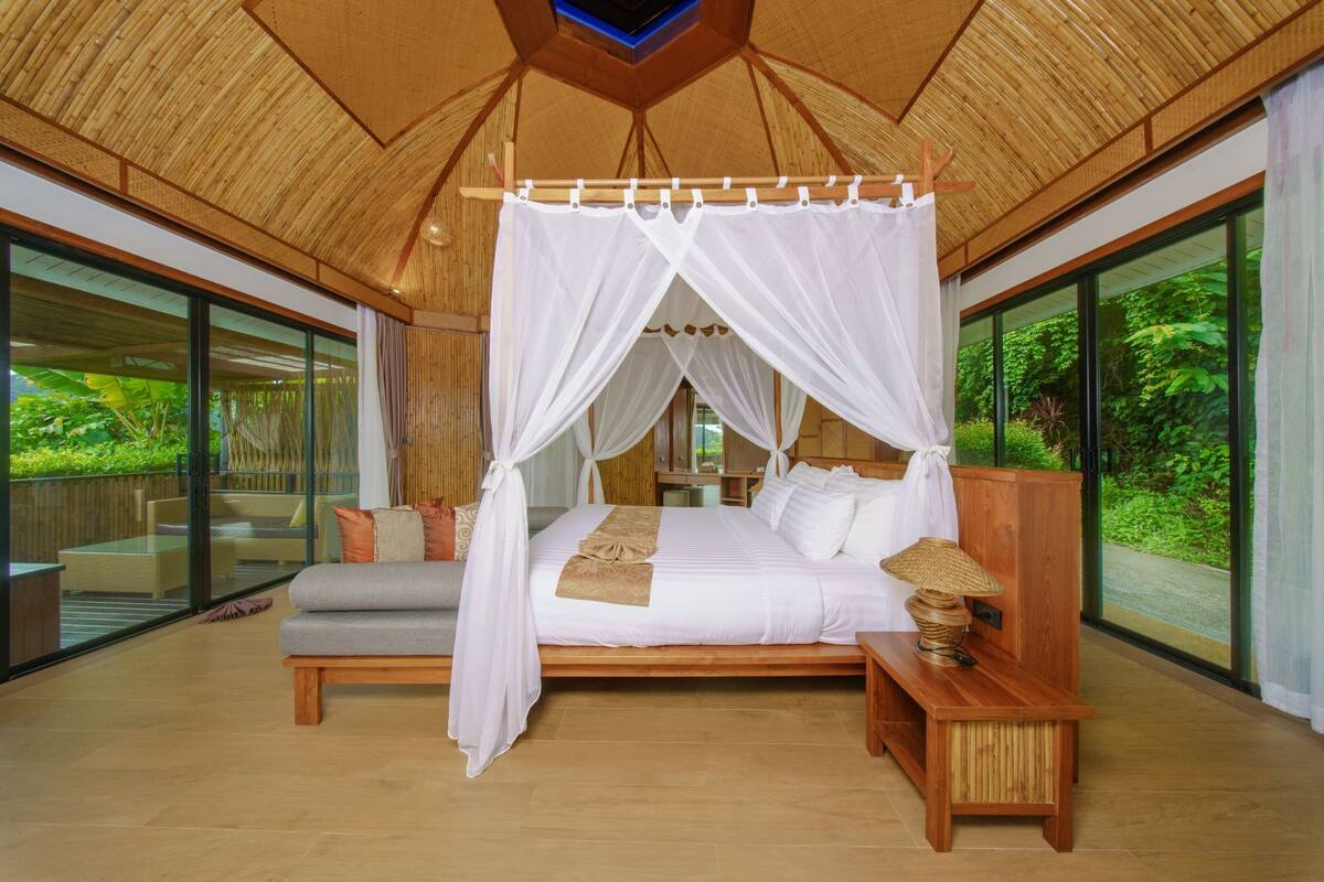 A spacious bedroom with a canopy bed, a wooden bench, and a view of lush greenery.