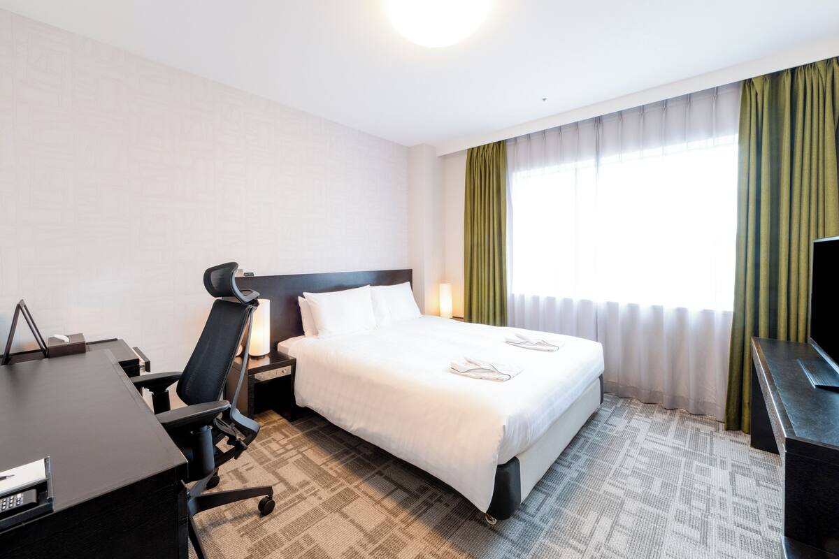 Una habitación de hotel con cama, escritorio con silla, televisión y ventana con cortinas.