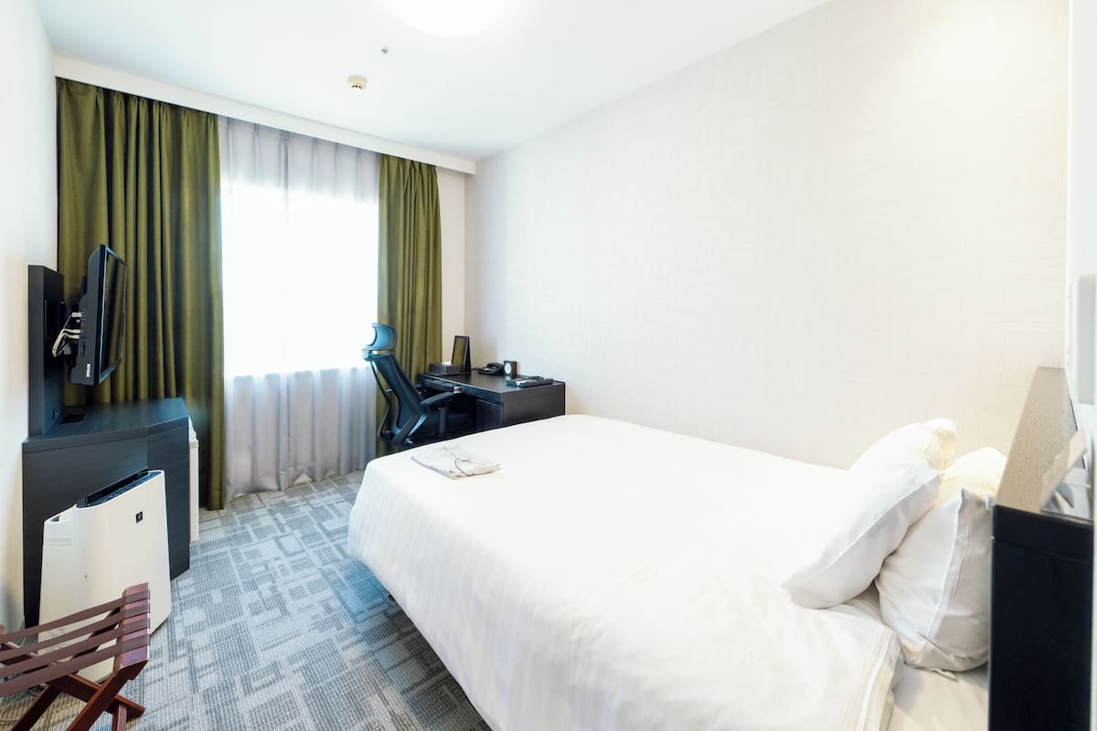 Una habitación de hotel con cama, escritorio con silla, televisor y ventana con cortinas.