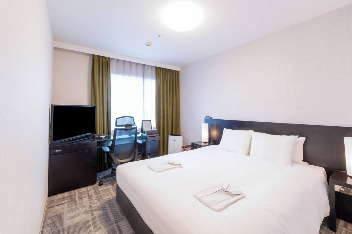 Habitación de hotel con una cama grande, un escritorio con computadora, un televisor y una ventana con cortinas.