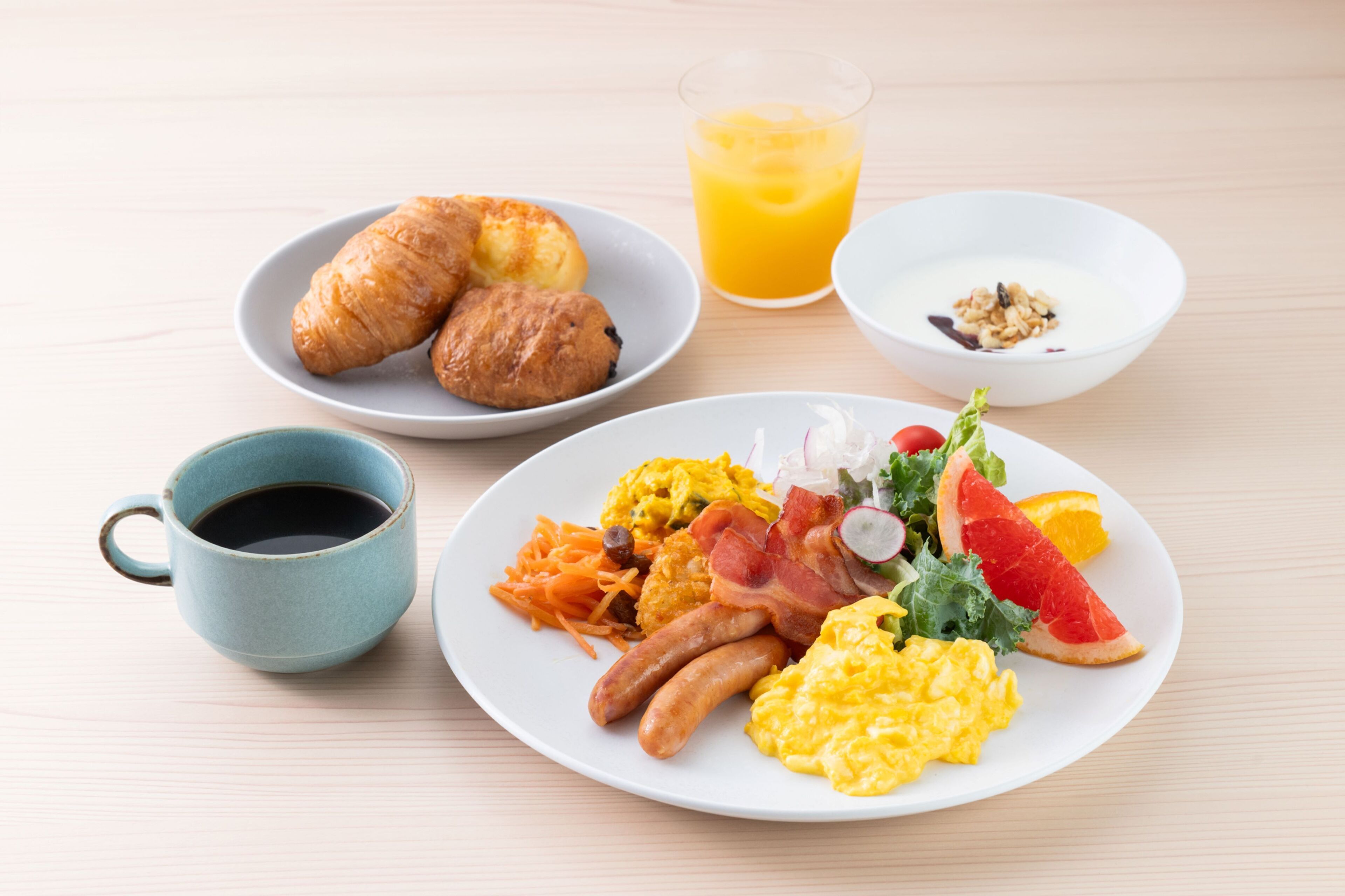 Un plato de desayuno con huevos, salchichas, tocino y verduras, un plato de croissants, una taza de café y un vaso de jugo de naranja.