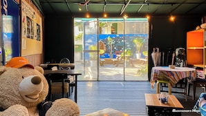 A cozy room with a large teddy bear, a table with a striped tablecloth, and a view of an outdoor area with a water feature and seating. - Backpacker's Home (Seogwipo)