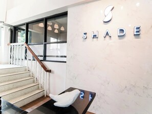 A staircase with a white railing, a wall with the word 'SHADE' in metallic letters, and a small table with a white chair. - Shade House - Bitec Bangna (Bangkok)