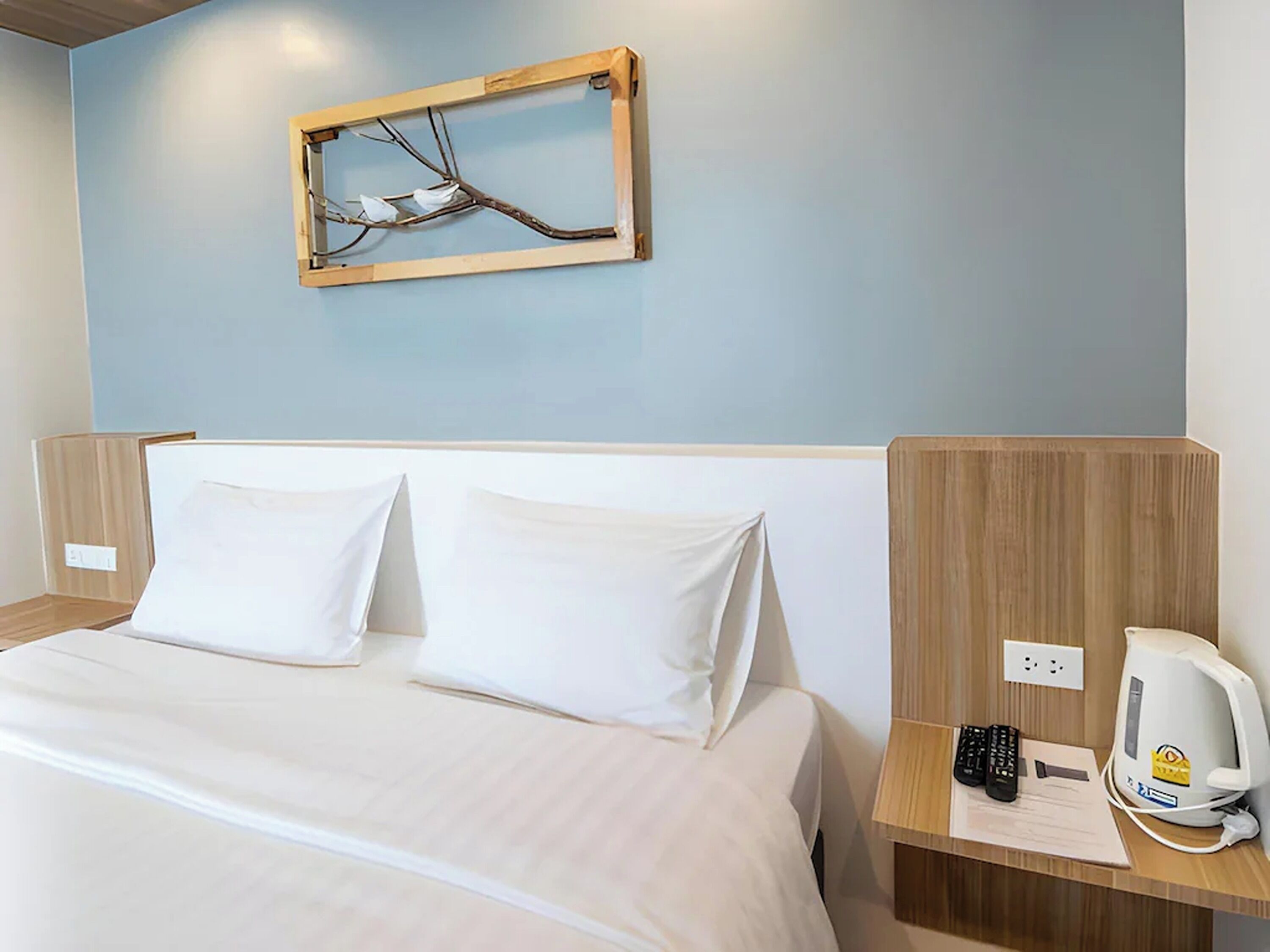A hotel room with a bed, two pillows, a wooden headboard, a nightstand with a phone, a kettle, and a wall-mounted picture frame.