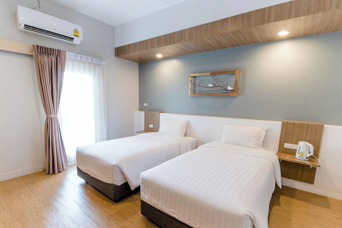 A hotel room with two beds, a wooden floor, a wall-mounted air conditioner, and a bedside table with a kettle.