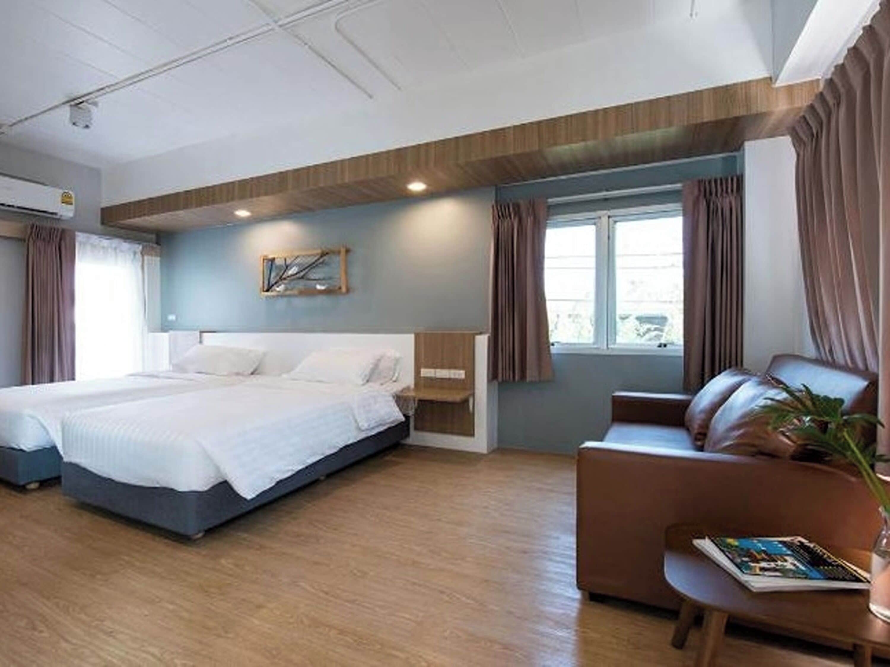 A modern hotel room with a large bed, a sofa, a small table with a magazine, and a window with curtains.