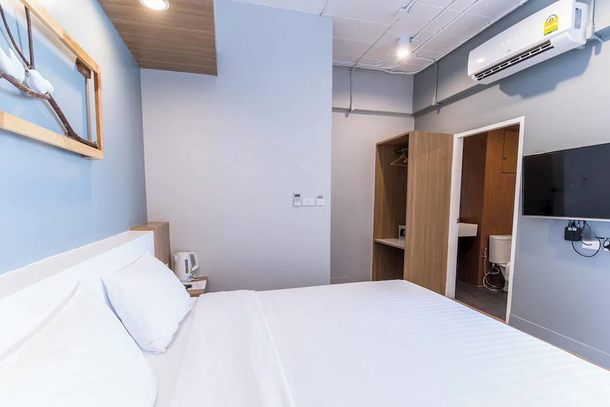 A hotel room with a bed, a bedside table with a kettle, a wardrobe, and an air conditioning unit.