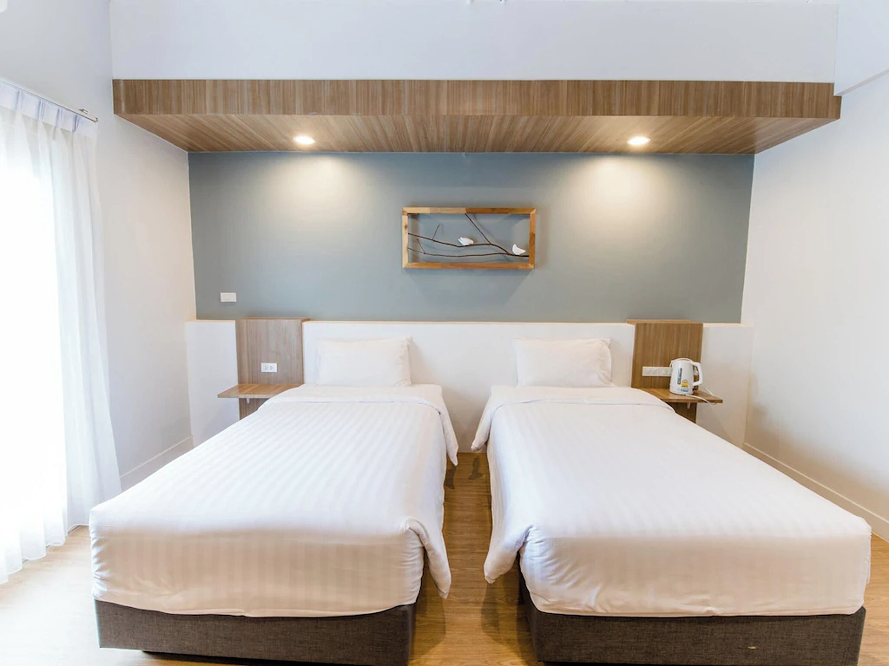 A hotel room with two single beds, a wooden ceiling, a wall-mounted light fixture, and a small table with a kettle.