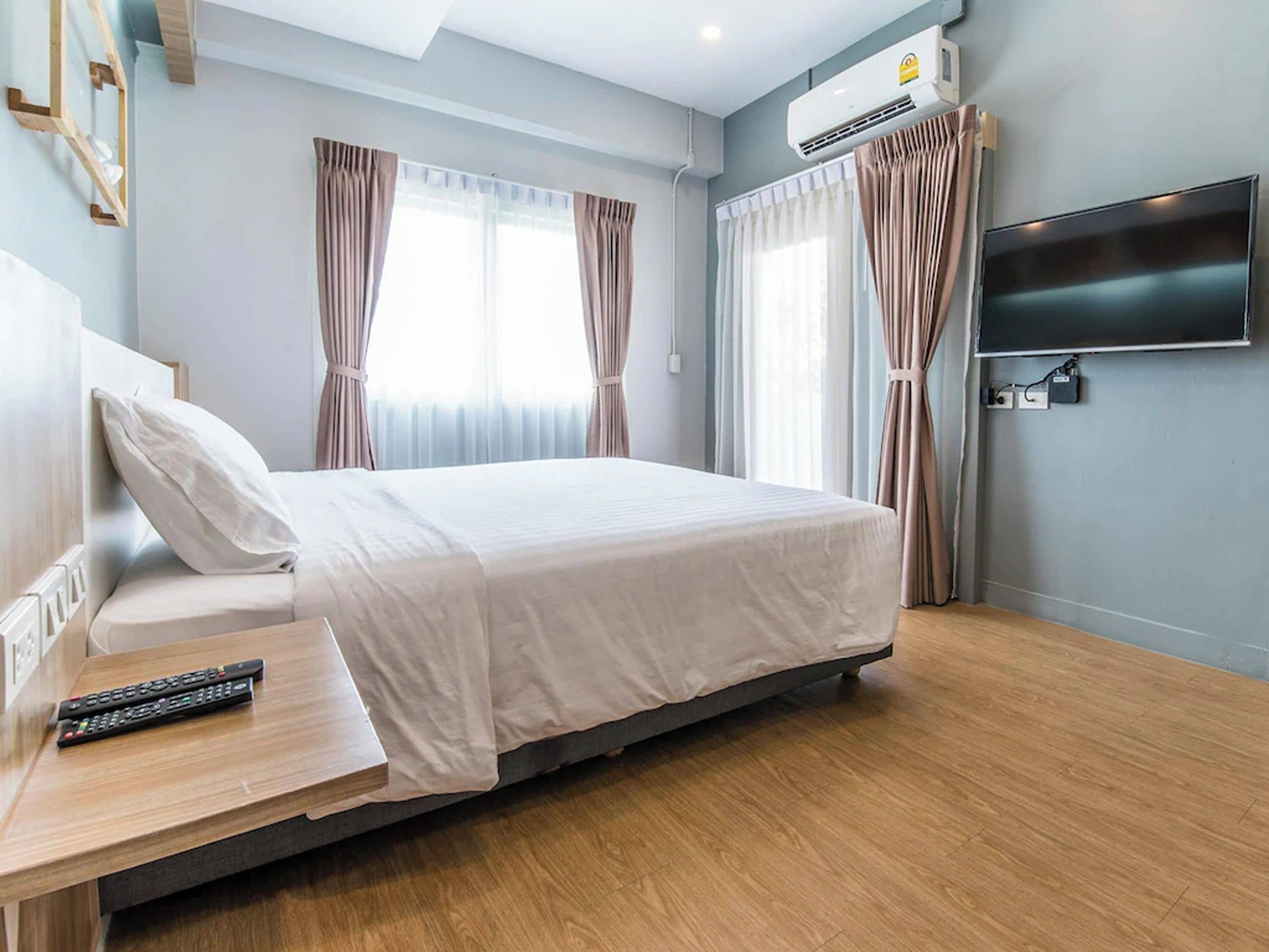 A hotel room with a bed, a TV mounted on the wall, an air conditioning unit, and a wooden desk with a remote control.
