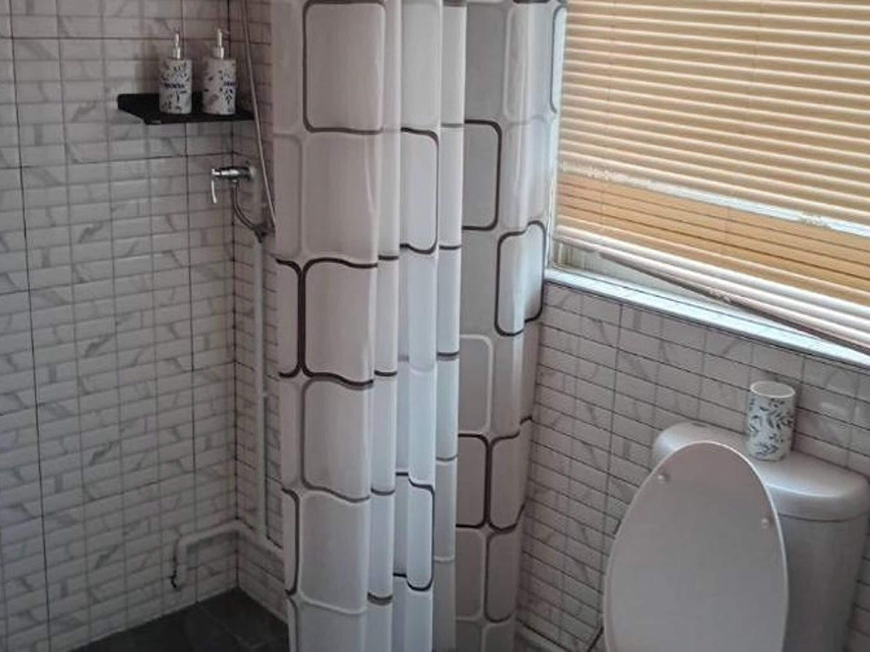 A bathroom with a tiled wall, a shower curtain, a toilet, and a window with blinds.