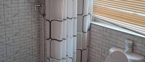 A bathroom with a tiled wall, a shower curtain, a toilet, and a window with blinds.