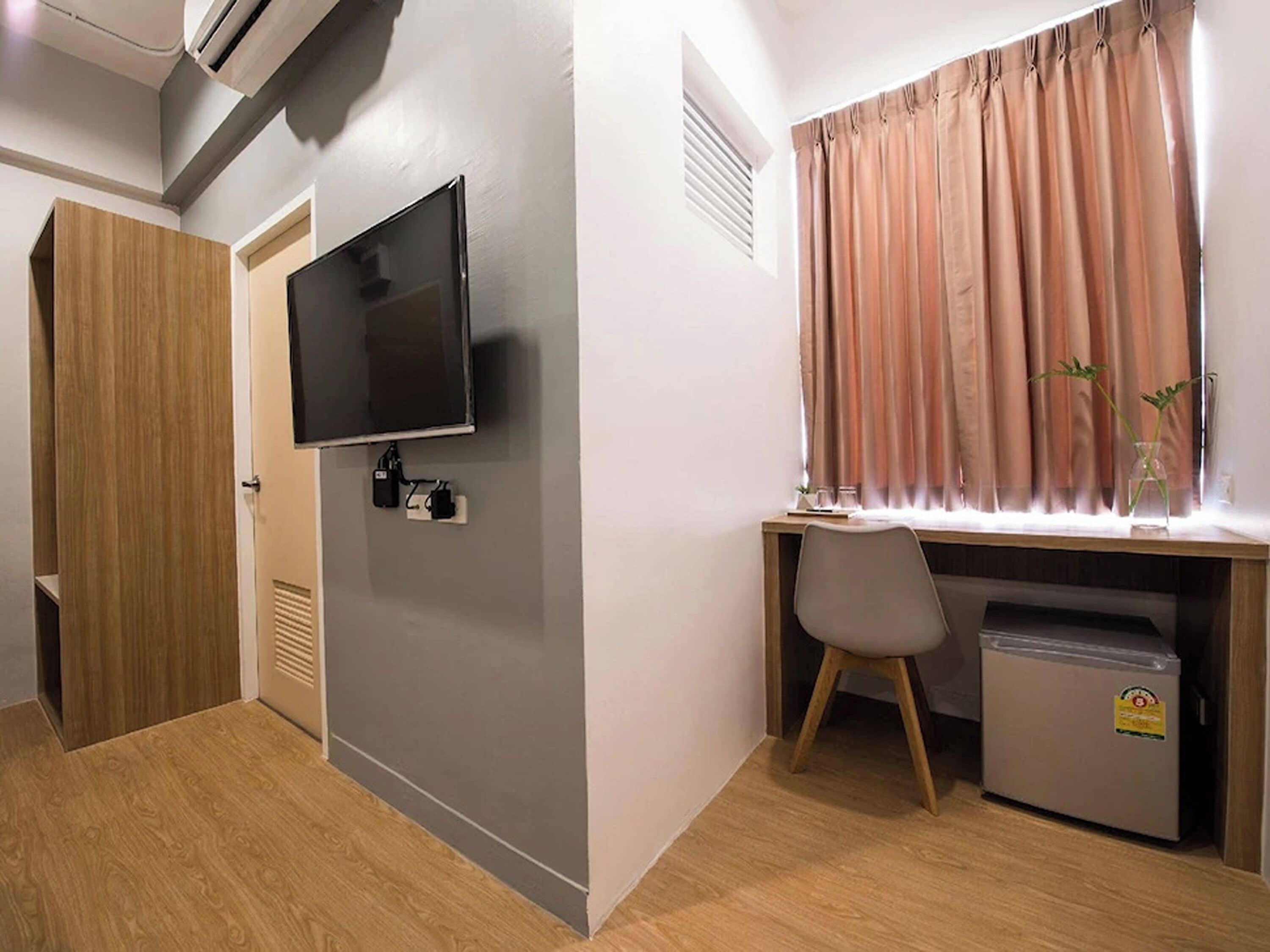 A compact room with a TV mounted on the wall, a desk with a chair, a small refrigerator, and a window with curtains.