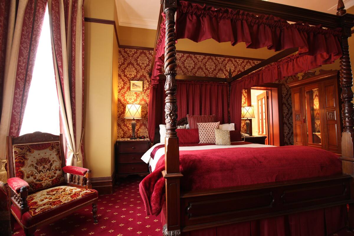 A four-poster canopy bed with a red bedspread, a wooden chair with a floral pattern, and a patterned carpet.