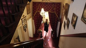 A grand staircase with a wooden handrail, leading to a room with red curtains and a chandelier. - The Grand On Macfie (Devonport)