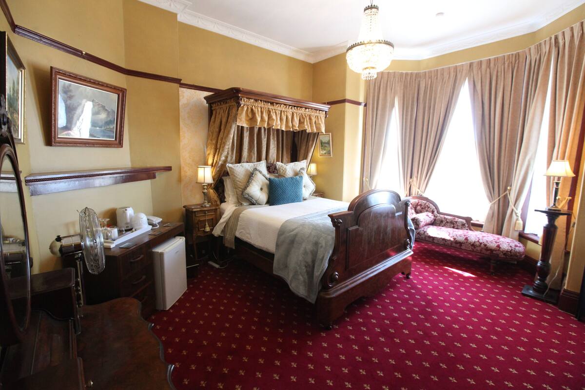 A traditional room with a four-poster bed, a red carpet, a sitting area with a chair, and a chandelier.