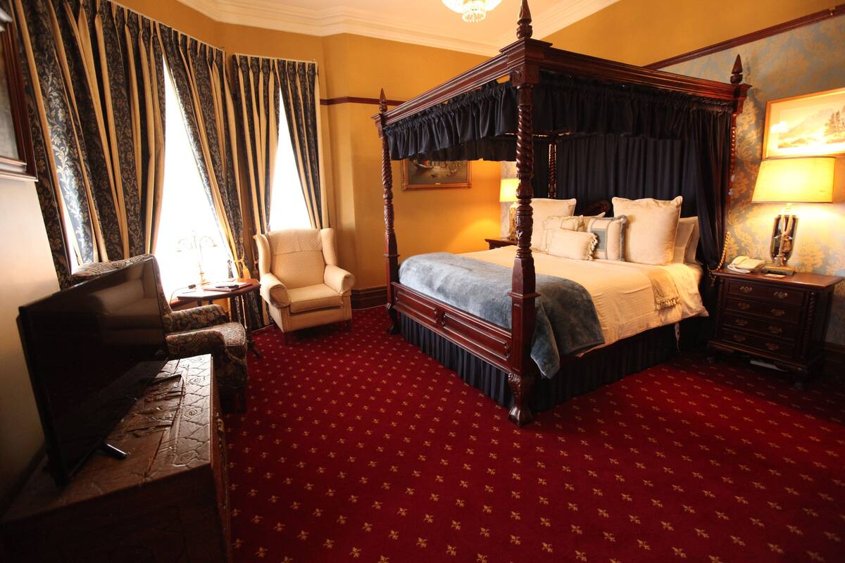 A four-poster bed with a red carpeted floor, a chair, a desk with a television, and a nightstand with a lamp.