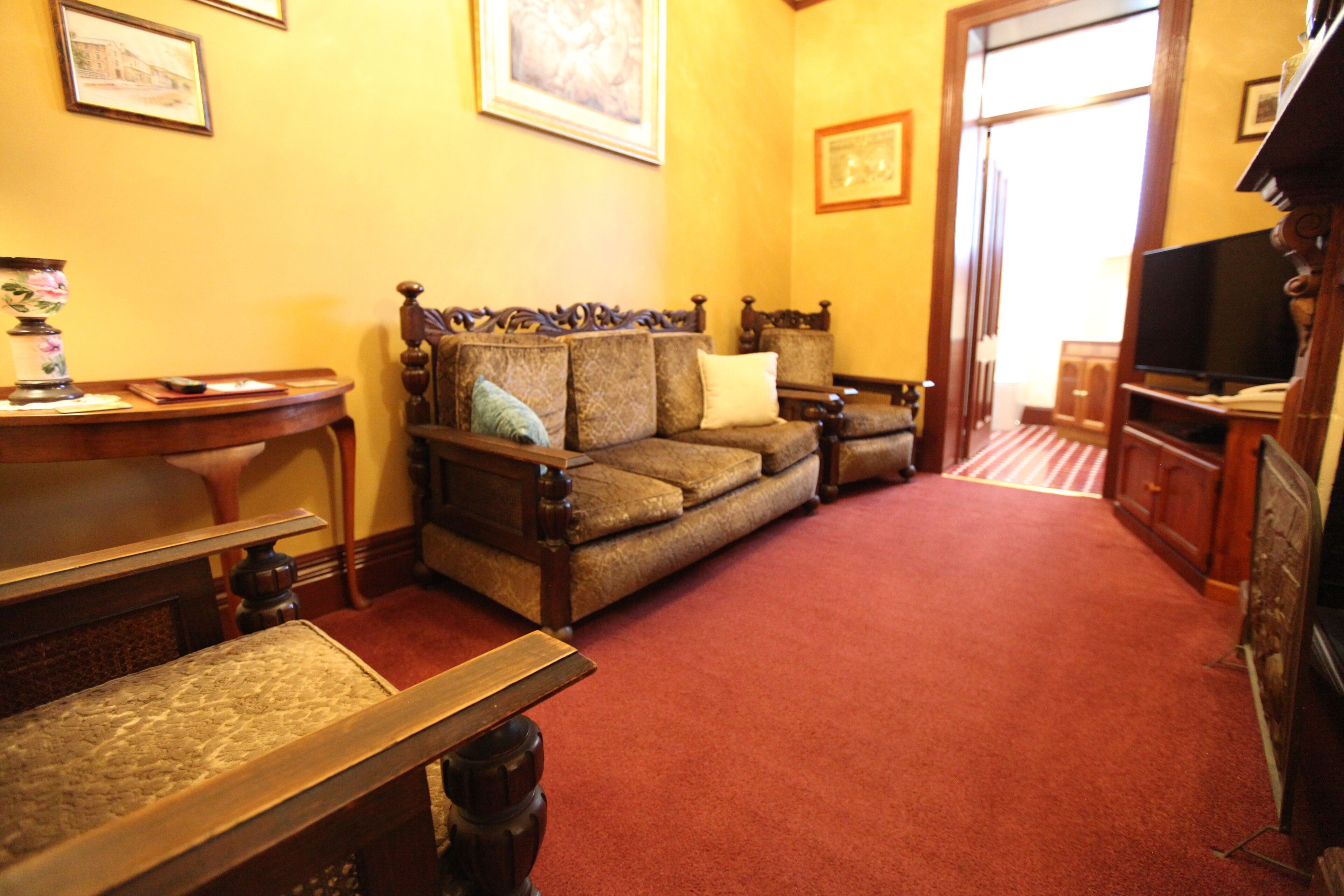 Una sala de estar con alfombra roja, un juego de sofá de madera, un televisor y una pequeña mesa con un jarrón y un libro.