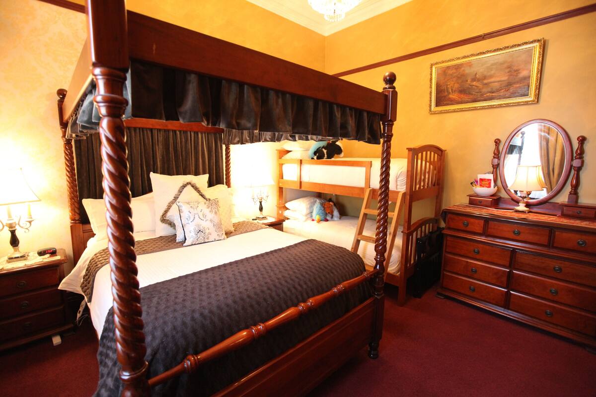 A four-poster bed with a dark headboard, a wooden dresser with drawers, a mirror, and a painting on the wall.