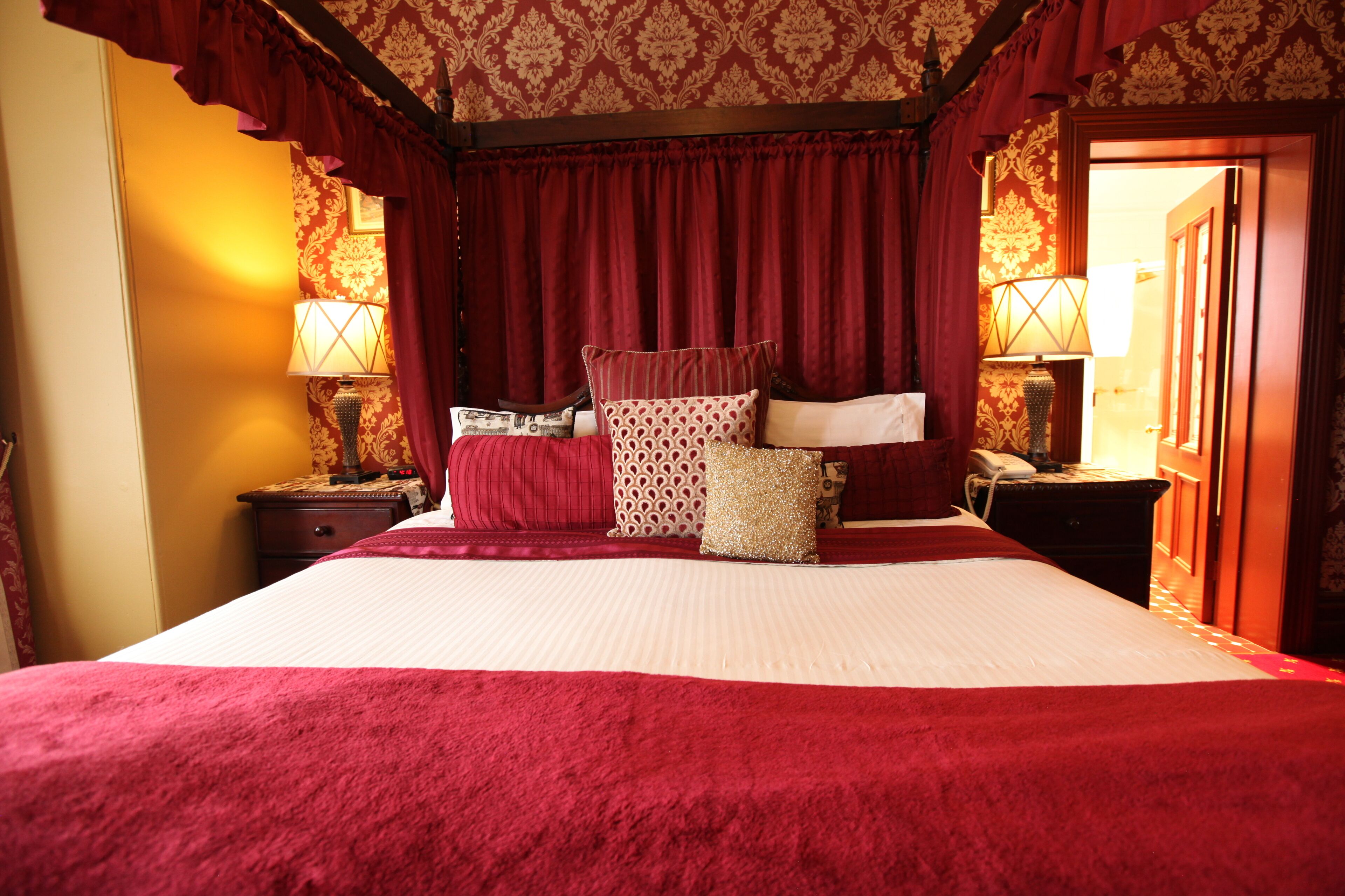 Una cama grande con una colcha roja, varios cojines y un cabecero de madera. Hay dos mesitas de noche con lámparas y un teléfono. La habitación tiene papel tapiz estampado y cortinas rojas.