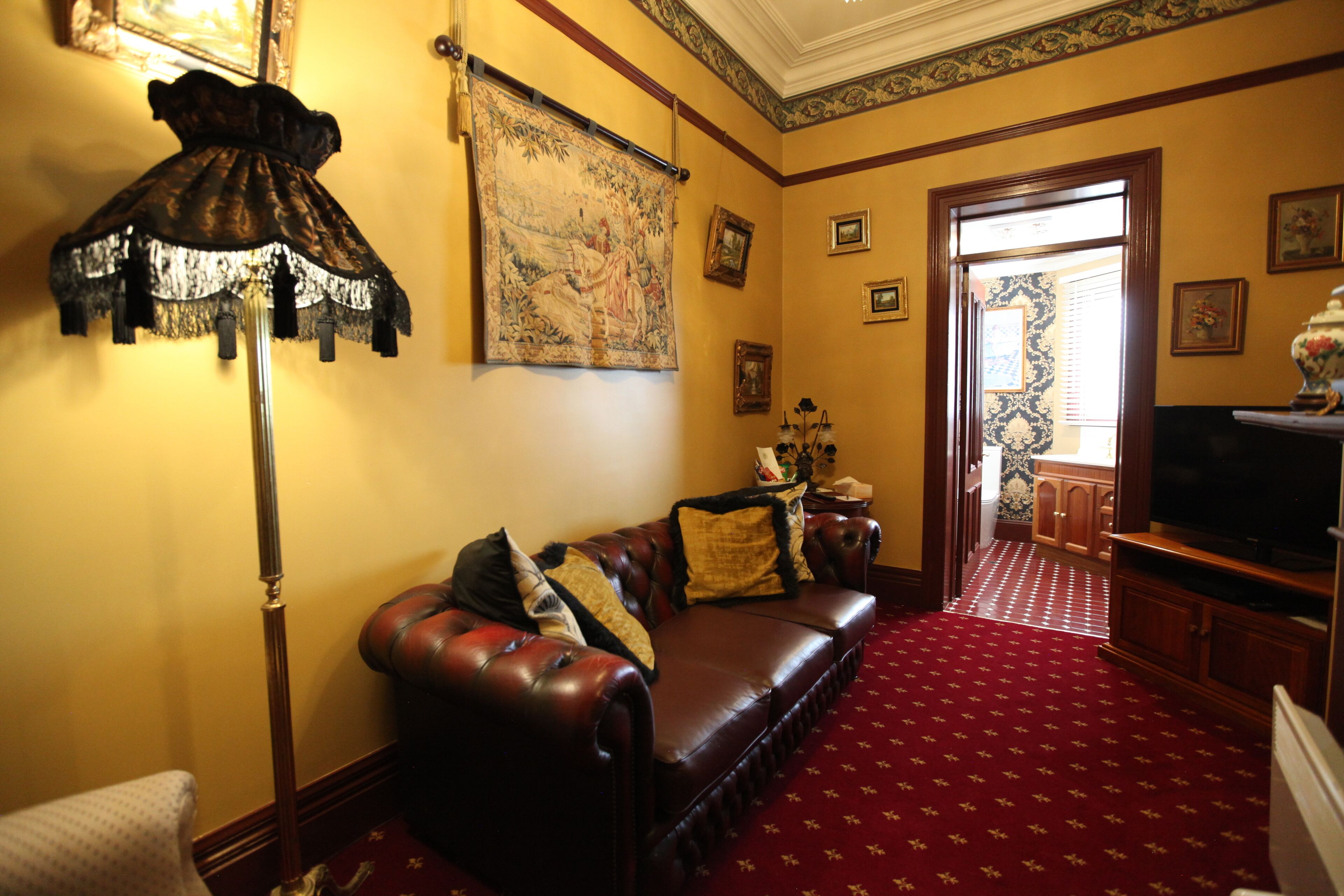 A living room with a red carpet, a dark brown leather sofa, a standing lamp, and a large tapestry on the wall.
