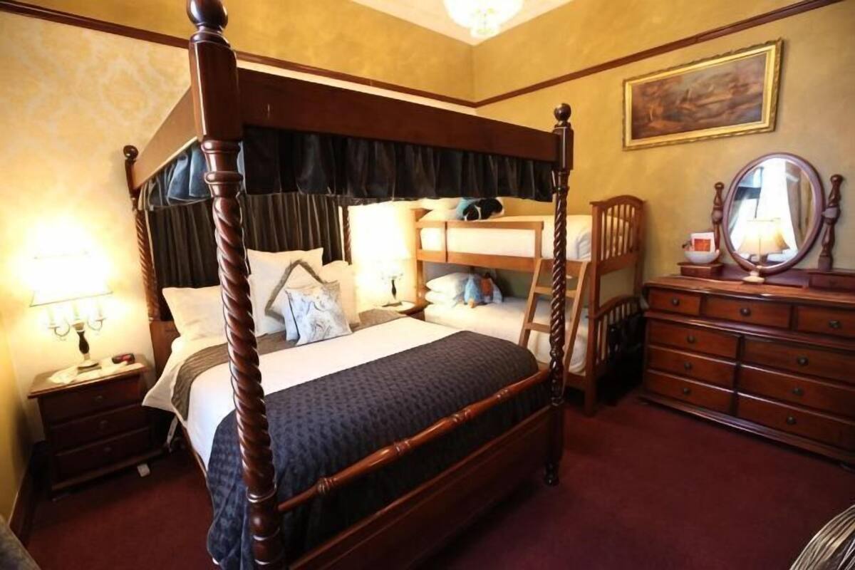 A four-poster bed with a dark headboard, a nightstand with a lamp, a dresser with a mirror, and a painting on the wall.