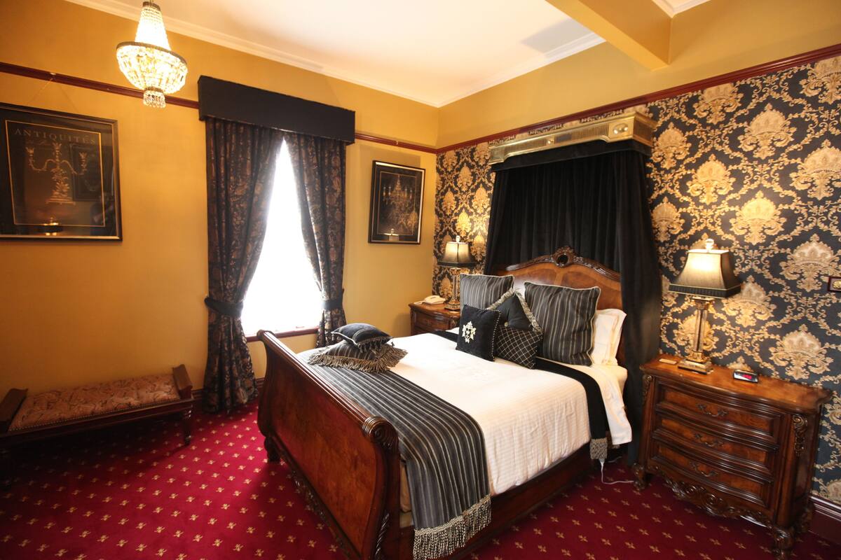 A bedroom with a large bed, a wooden nightstand, a dresser, a chandelier, and patterned wallpaper.