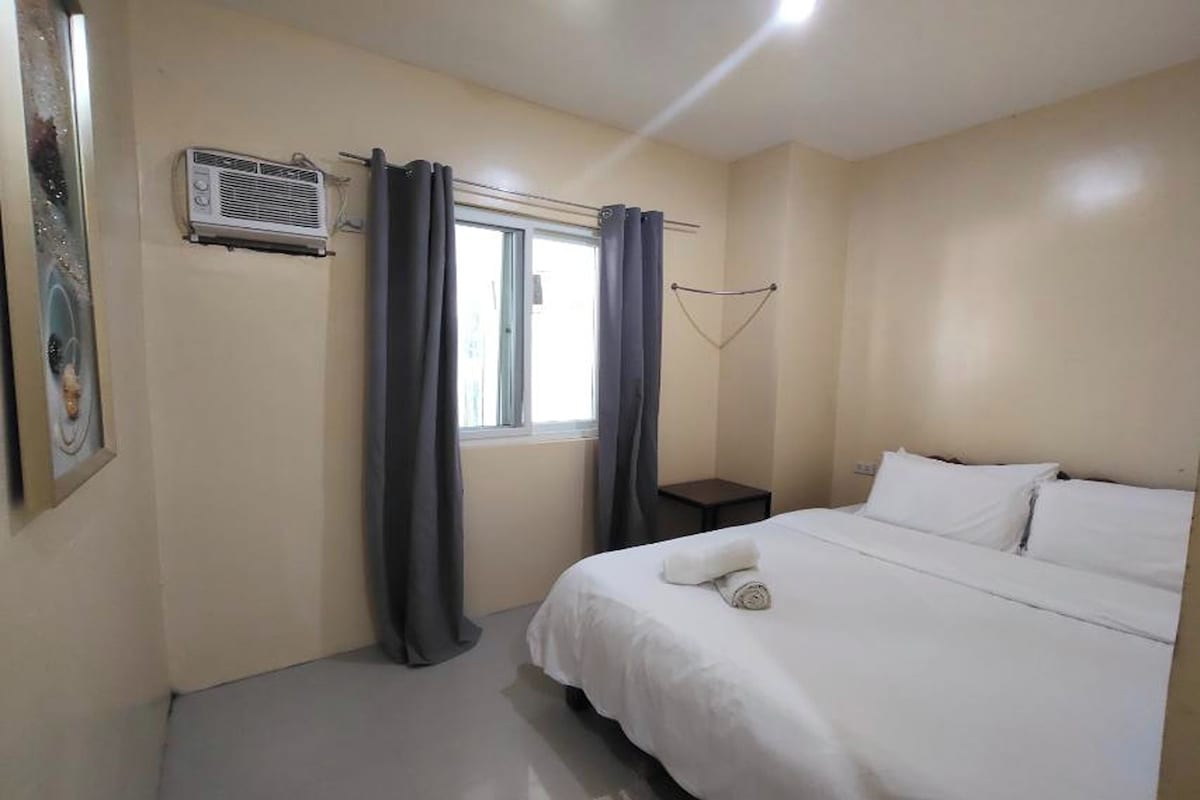 A bedroom with a bed, a window with curtains, a small bedside table, and an air conditioning unit.