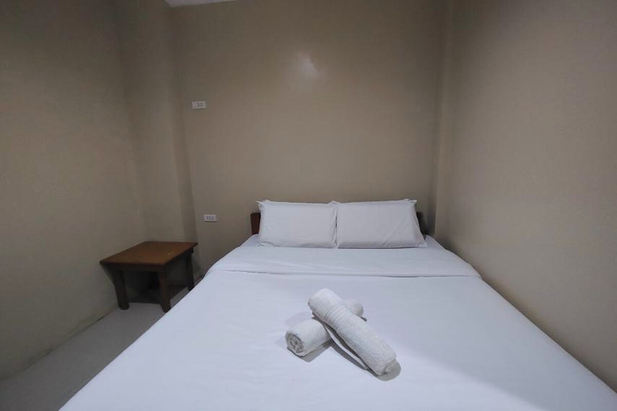 A small, single-bed room with a wooden nightstand and a rolled towel on the bed.