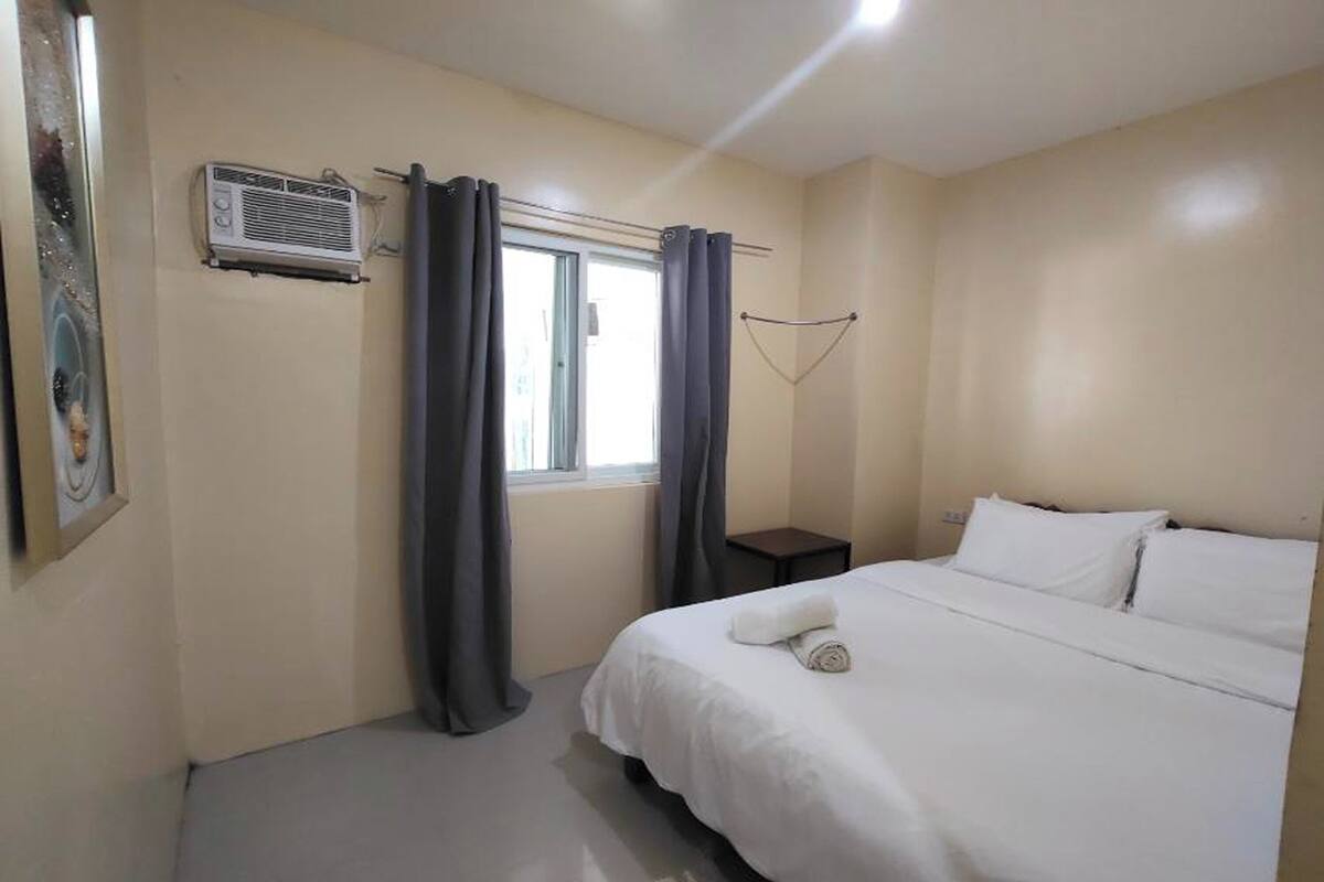 A bedroom with a bed, a window with curtains, a small bedside table, and an air conditioning unit.