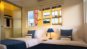 A hotel room with two beds, a window with a view of buildings, a bedside table with a lamp, and a small shelf with towels. - The Residence (Colombo)