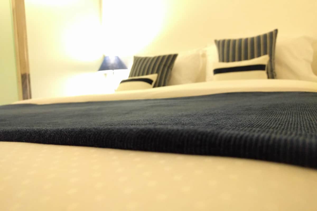 A bed with a dark blue blanket and white pillows, a lamp on the nightstand, and a window with curtains.
