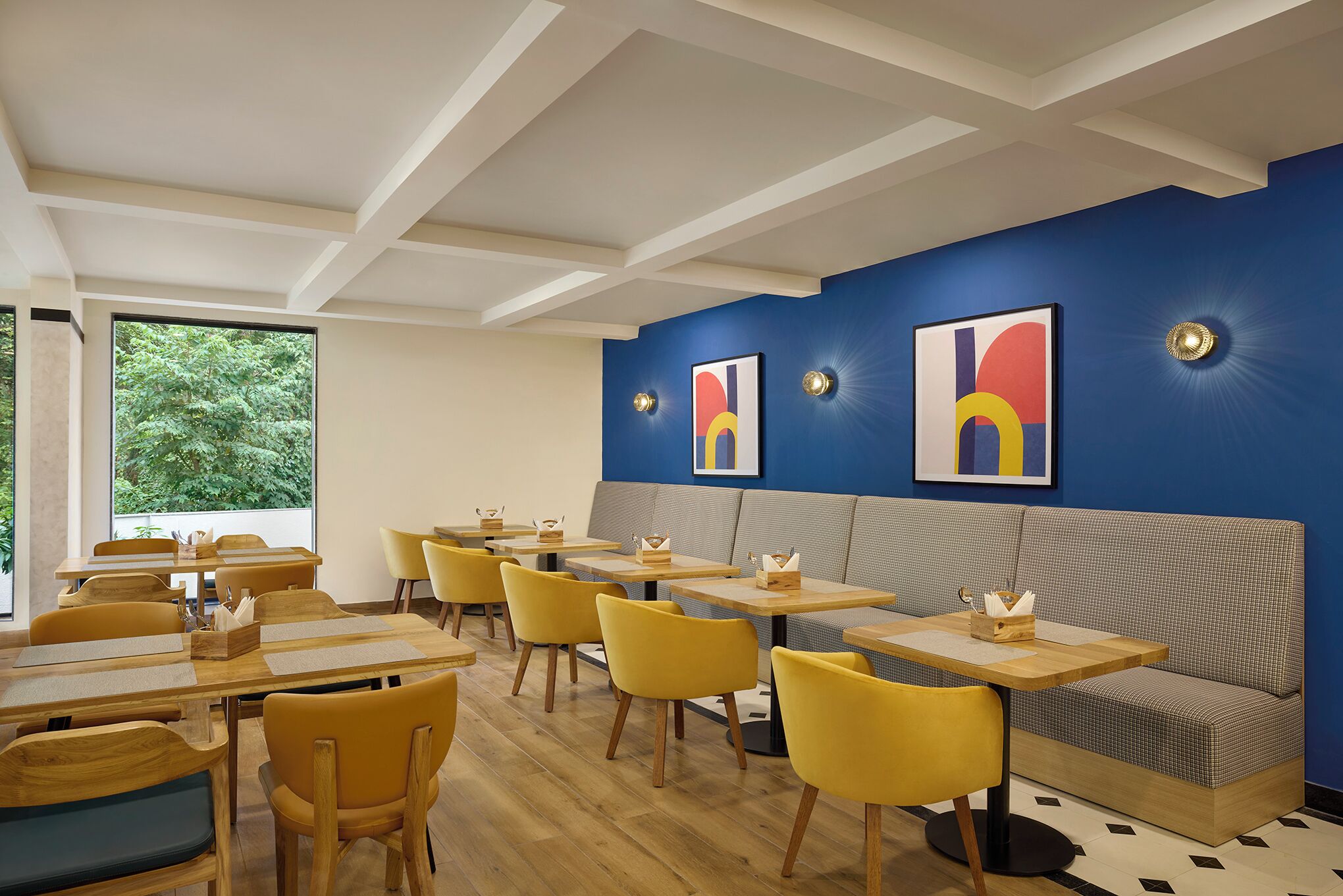 A modern dining area with wooden tables and yellow chairs, a blue accent wall, and artwork.