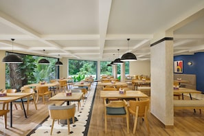 Un vaste espace de restauration intérieur, doté de nombreuses tables et chaises, de grandes fenêtres offrant une vue sur la verdure et d’un éclairage suspendu.