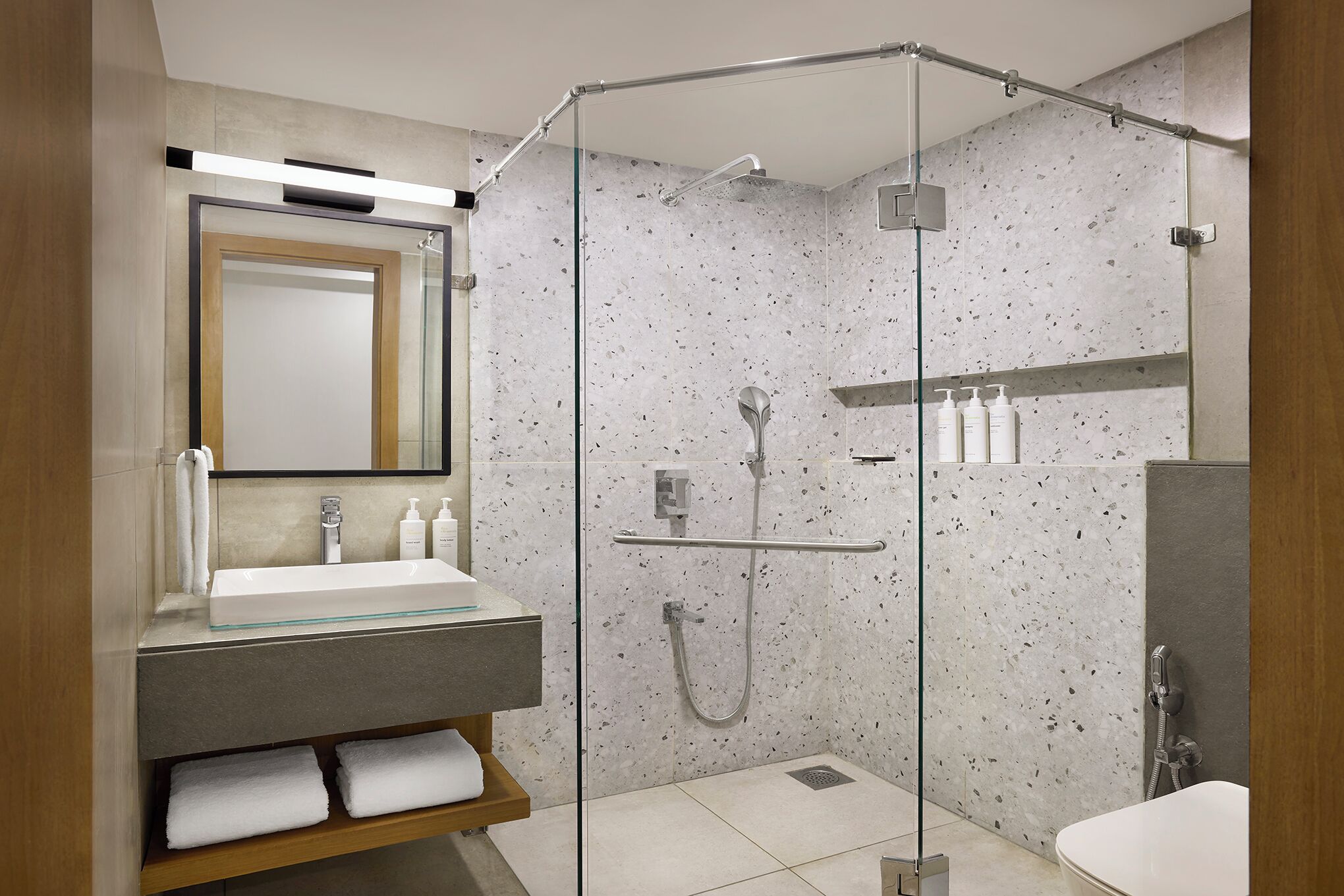 Un baño moderno con ducha envuelta en vidrio, un espejo grande y un tocador con lavamanos y espacio para toallas.