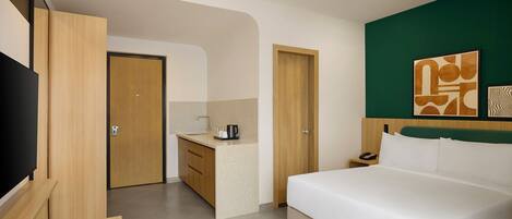 A hotel room with a bed, a desk, a chair, a kitchenette, and artwork on the wall.