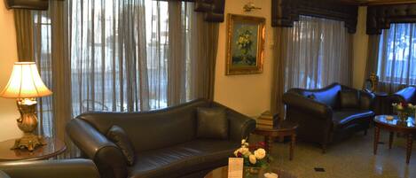 Una sala de estar con un sofá de cuero, una mesa de centro y una lámpara. Hay un cuadro en la pared y una ventana con cortinas.