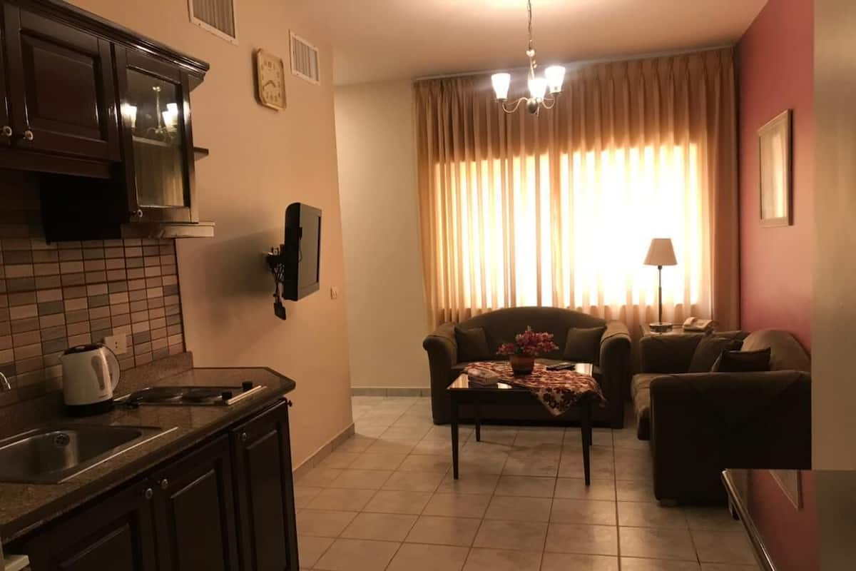 A kitchen with a coffee maker, a dining table with a floral tablecloth, and a sofa in a living area with curtains.