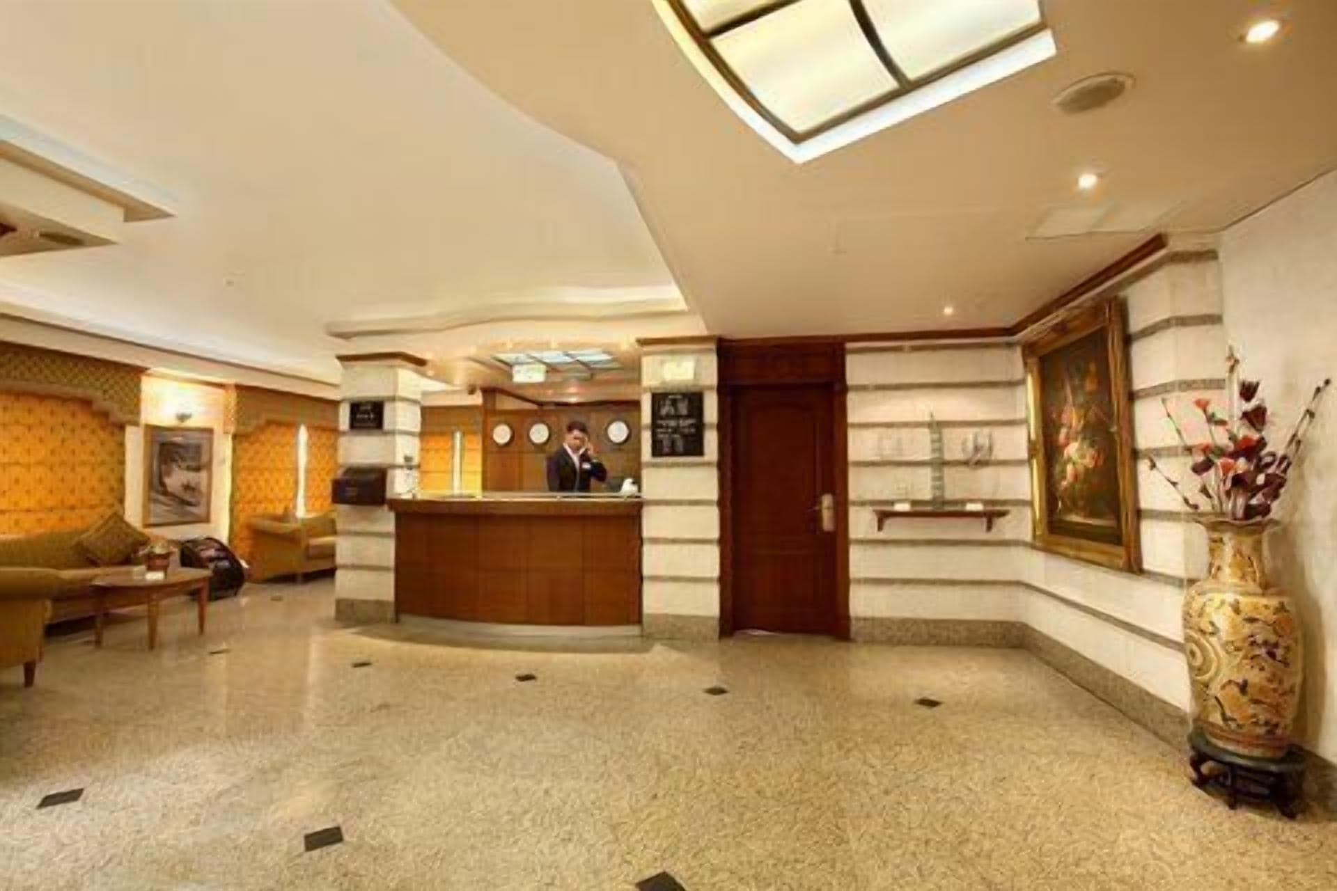 Una hall d'albergo con una reception, un divano, un tavolino, una sveglia e un vaso con fiori.