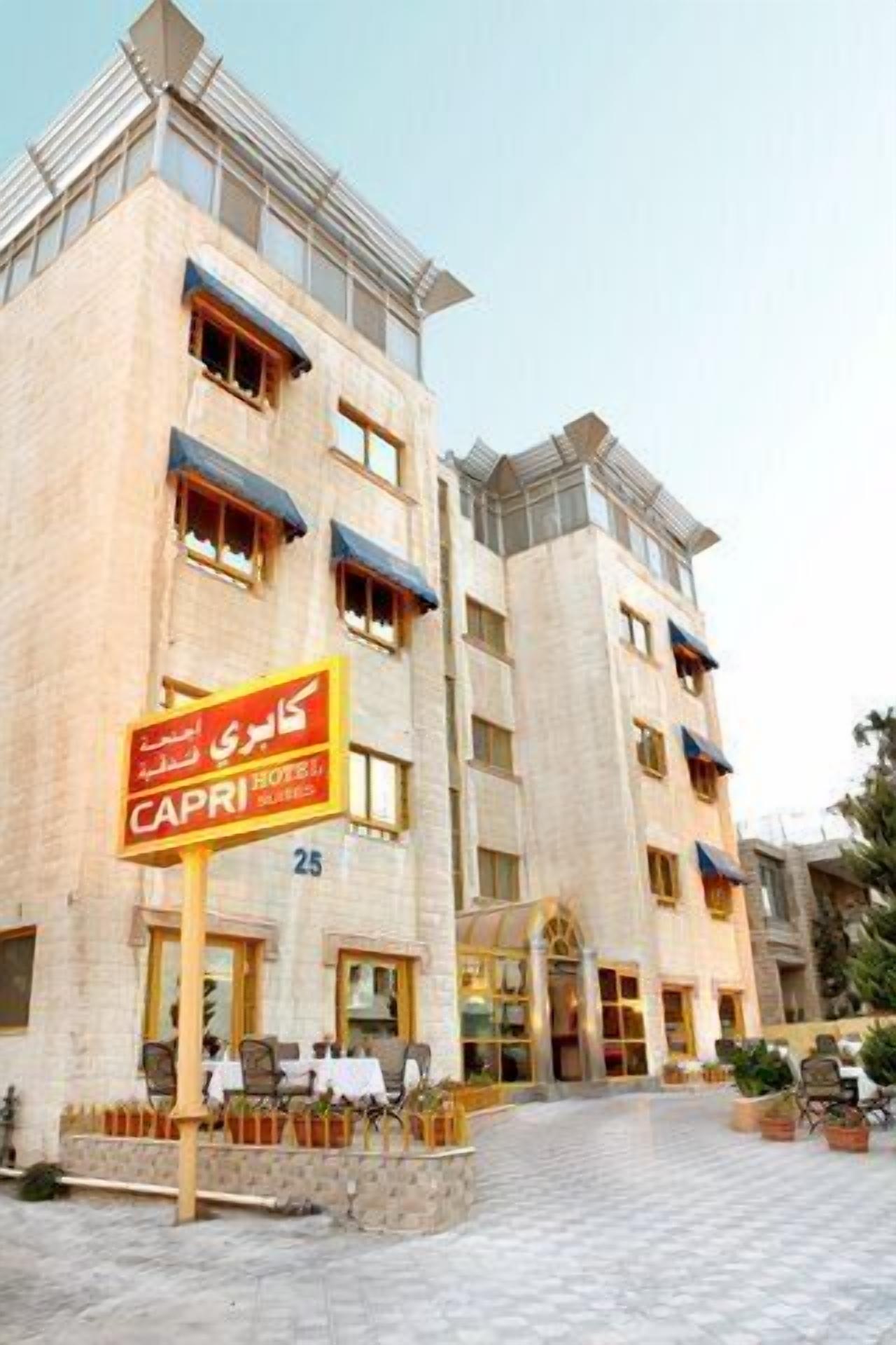 A hotel named "Capri" with a sign in a foreign language, multiple balconies, and a paved courtyard with outdoor seating.