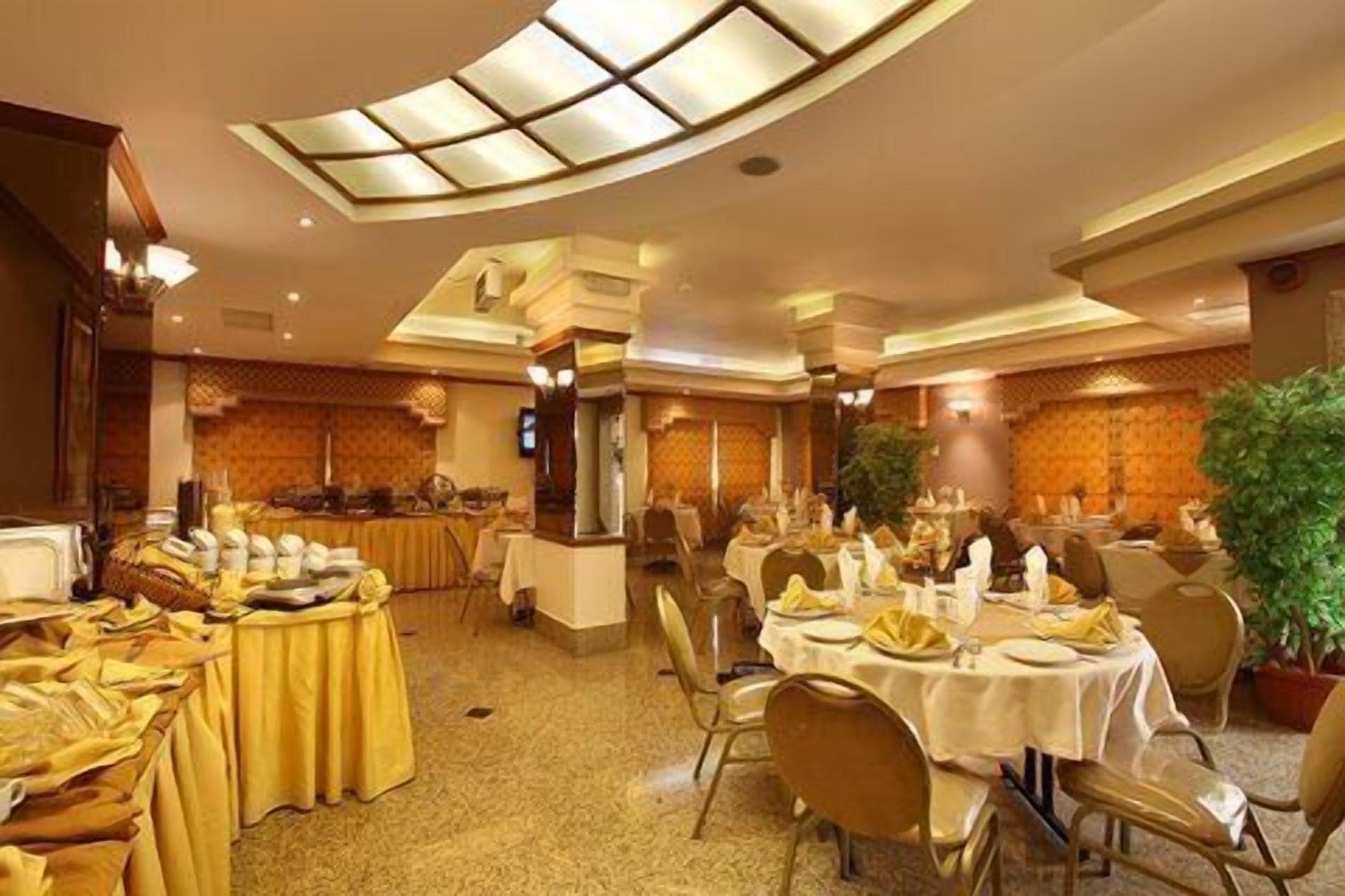 A banquet hall with round tables set for dining, a central buffet area, and a large potted plant.