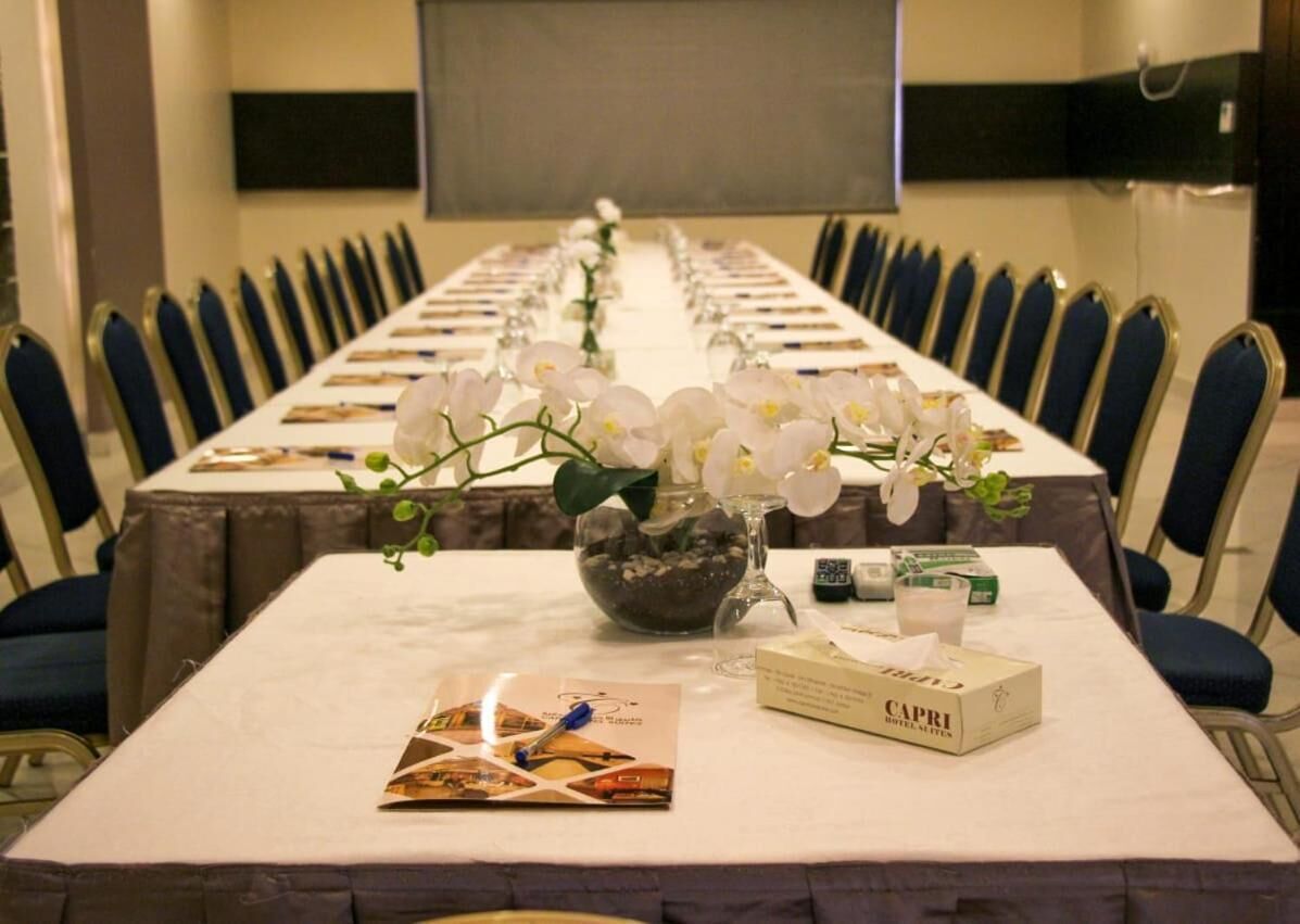 Un salón de conferencias con una larga mesa, sillas, una pantalla de proyector y un jarrón con flores blancas.